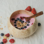 Natural white coconut bowl filled with a vibrant smoothie topped with fresh strawberries, blueberries, raspberries, banana slices, and granola, accompanied by a wooden spoon on a light-coloured background